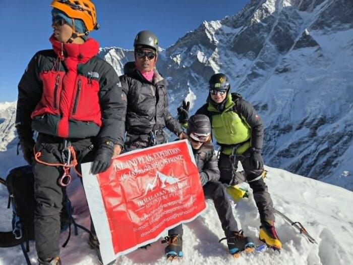 Several people on a summit with a higher peak behind. 