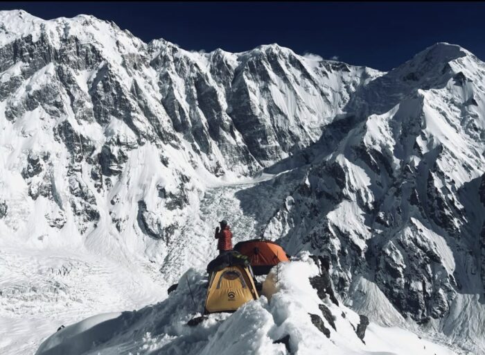A pereson standings in front of two tens on a snowy ridge, with a big mountain face in background. 