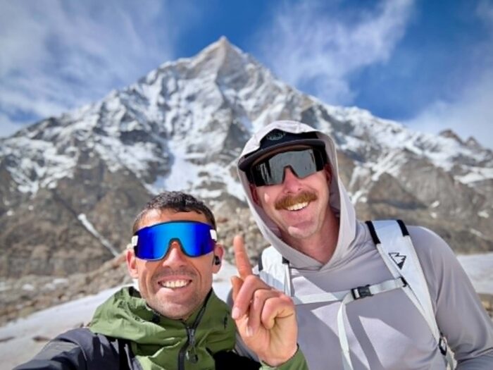 Two climbers smile with a peak behind them. 