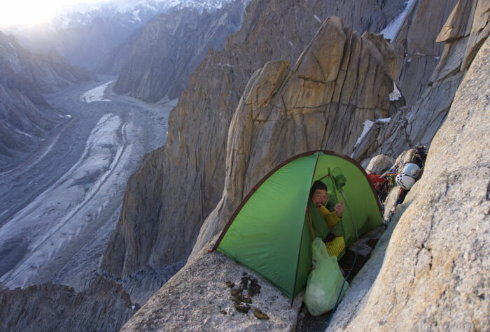 A climber in a small tent set on a rocky ledge facing the void.