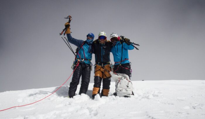 Three climbers on a flat, snowy summit, one rising his ice axe.