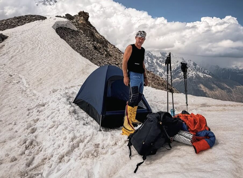 A climber in short sleves by a tent on a snowy ridge.