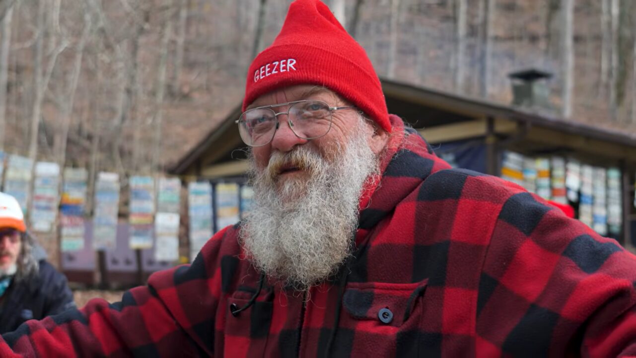 An old man standing by a cabin in the woods