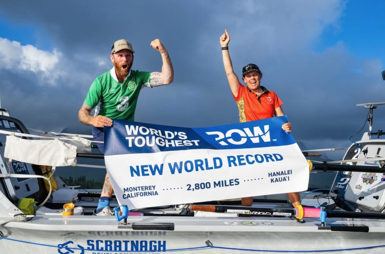 OceanWays take first place in the World's Toughest Row, and stand aboard their boat at the finish line holding their speed record banner.