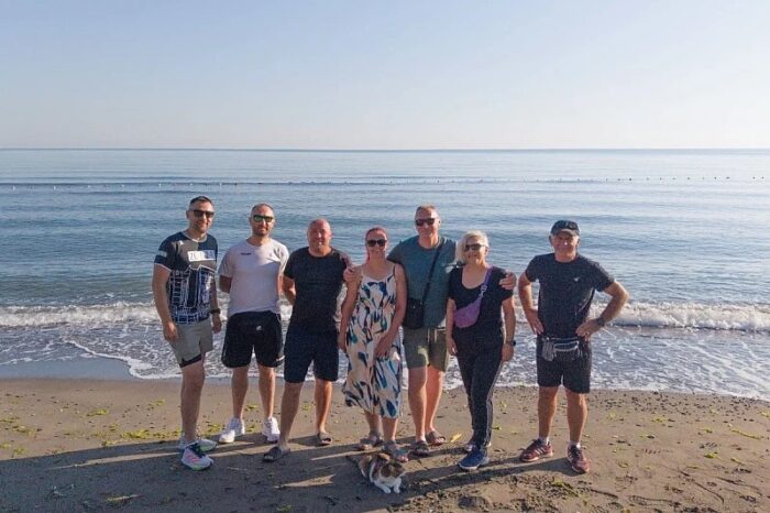 A group of people pose on the shore of the sea, with a cat.