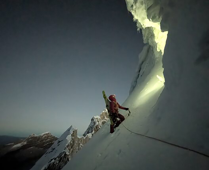 A climber ends a traverse on snow and looks at an ice cave lit up by a partner's headlamp.