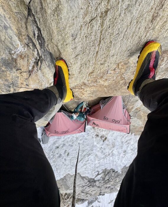 Two portaledges between a steep snow ramp and a vertical wall,a s seen between the legs of a climber.