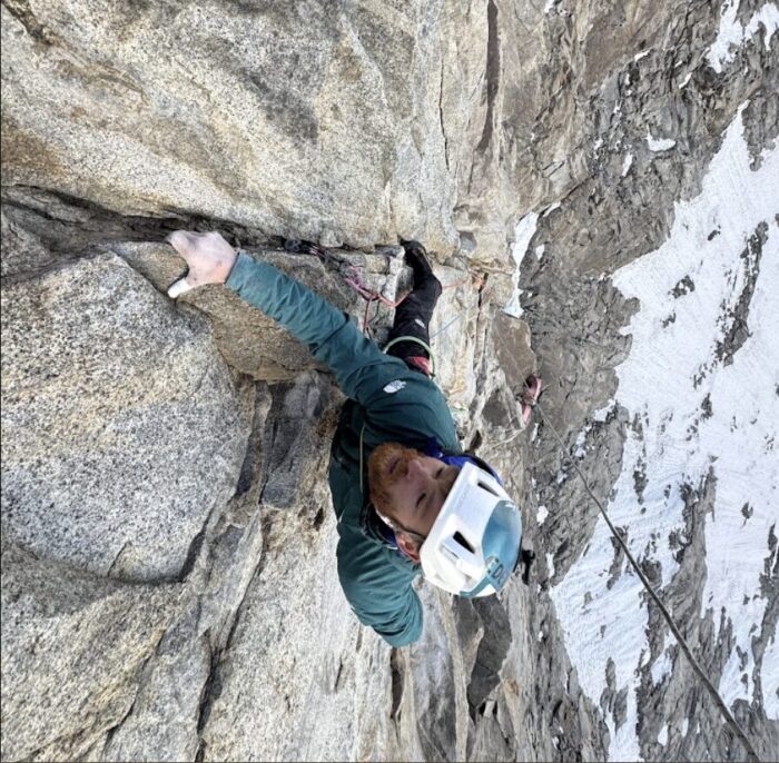 A climber on a vertical rock face, snow down below him.