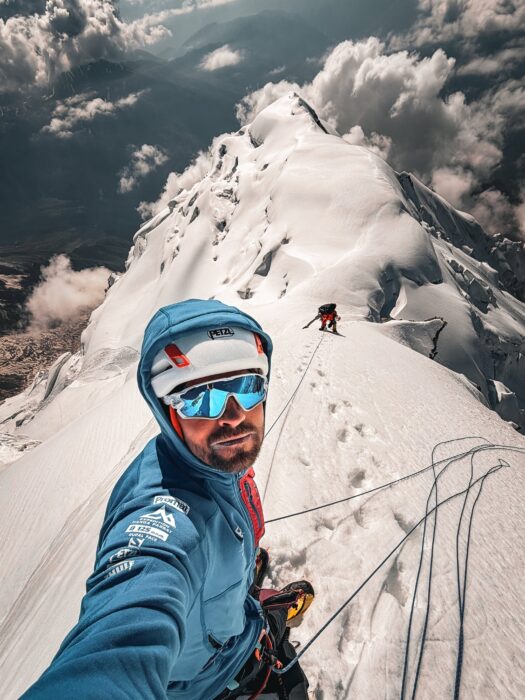 Two climbers roped up on a sharp snow ridge, the one in front taking a selfie. 