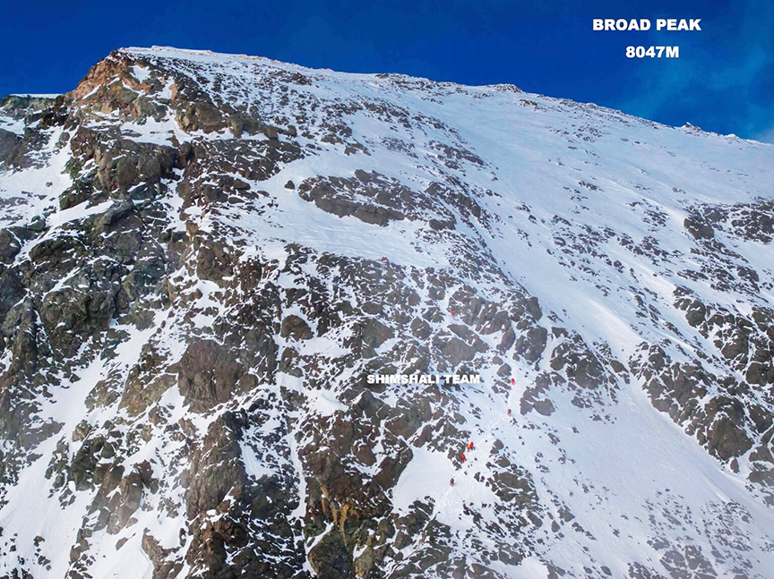 Climbers look tiny as they go up in a line up a big mountain face.