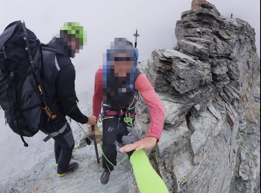 A climber with pixelated face pushes the person holding the camera on a rocky ridge.