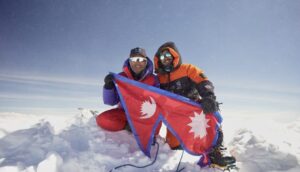 Two Nepalse climbers on a snowy summit holding a Nepali flag.