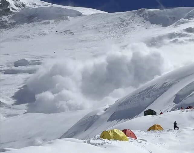 An avalanche falls down a snowy slope to the left of a group of tents.