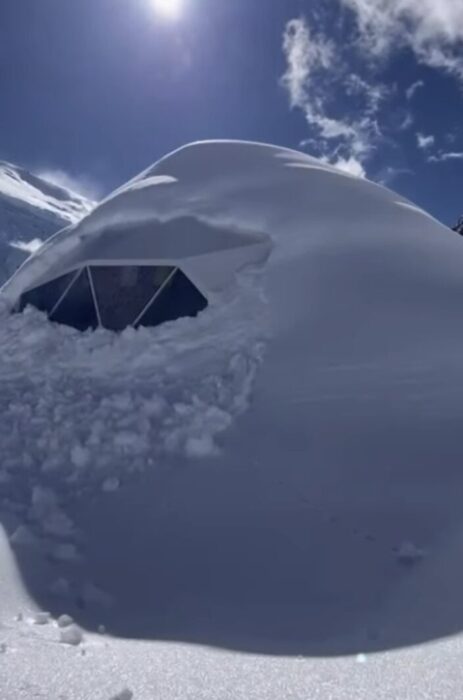 Dome tent buried in snow, with only a n edge of the window visible.