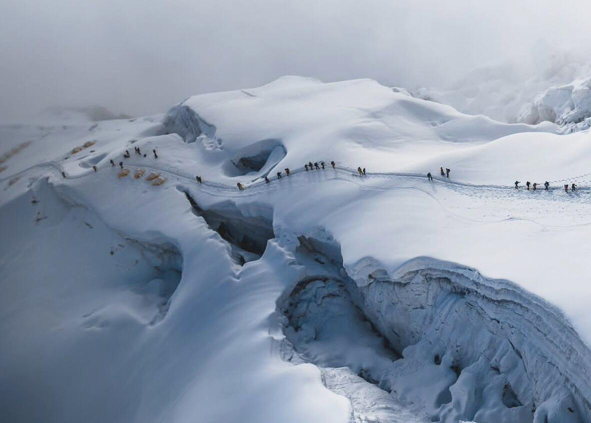 A line of climbers follow a trail in the snow of Manaslu, with tents on the left and big crevasses below them.