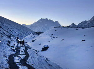 Trekkers with headlamps still on at dawn as they walk on a snowy plain toward the mountains.