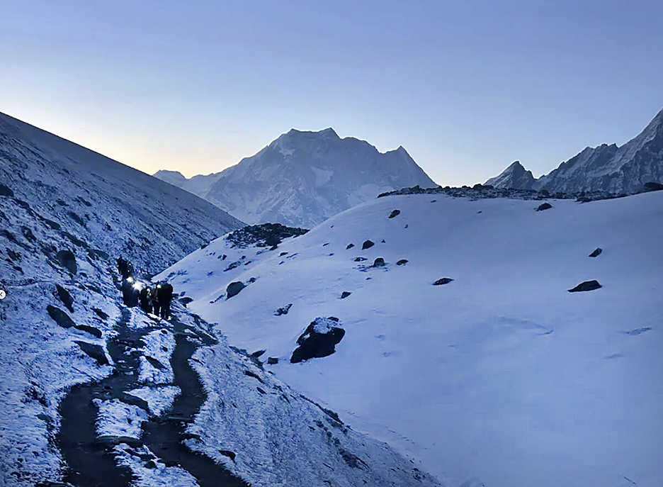 Trekkers with headlamps still on at dawn as they walk on a snowy plain toward the mountains.