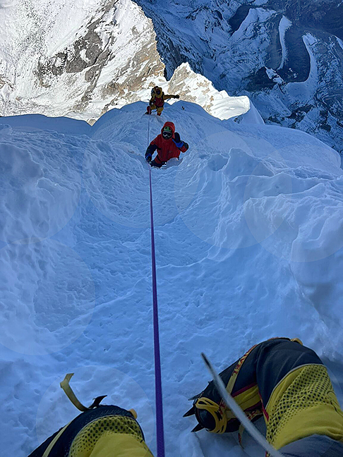 Climbers clipped to a fixed rope on a deep trail in a snowy, sharp ridge.