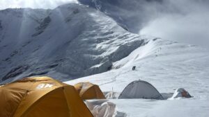 Tents on a big plane in front of Dhaullagiri, and some climbers following an uphill trail in the snow.