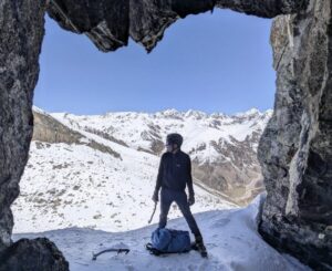A climber at the entrance of a mountain cave, peaks in background.
