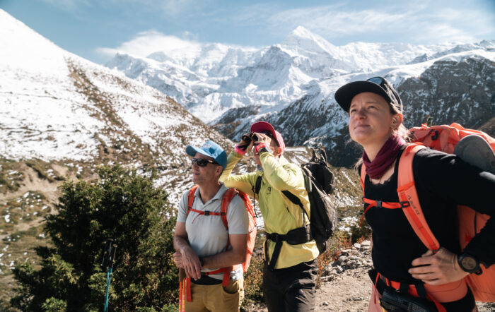 Three climbers look at mountains in front of them, one of them with binoculars, in a stunning mountain setting. 