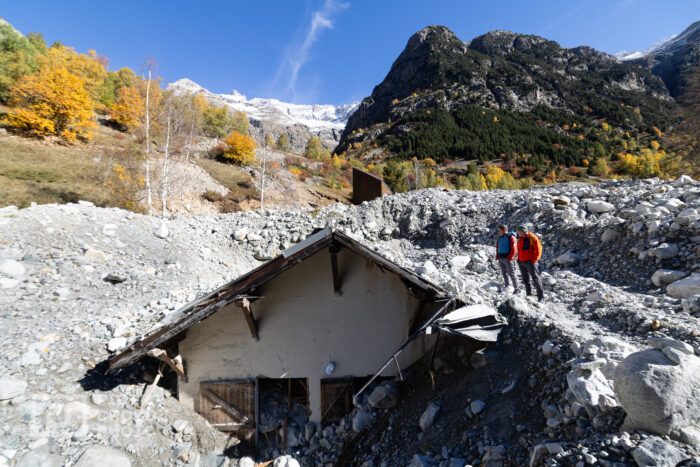 Two climbers watch a stone house semi-buried in debris. 