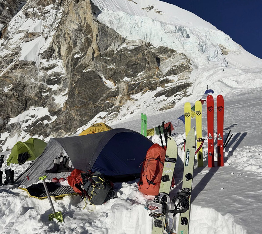 A tent and skis stuck in the snow in front of a himalayan face.