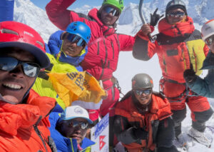 Sherpas in colorful clothes smile and raise hands on the summit of Ama Dablam in the sun.