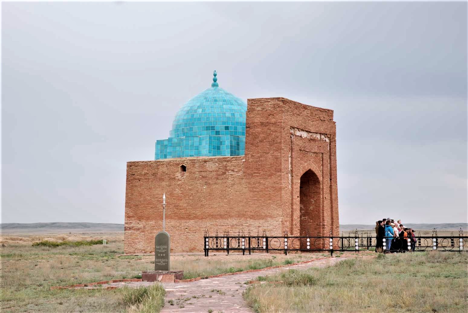 A square mausoleum made of reddish stone with a turquoise dome on top.